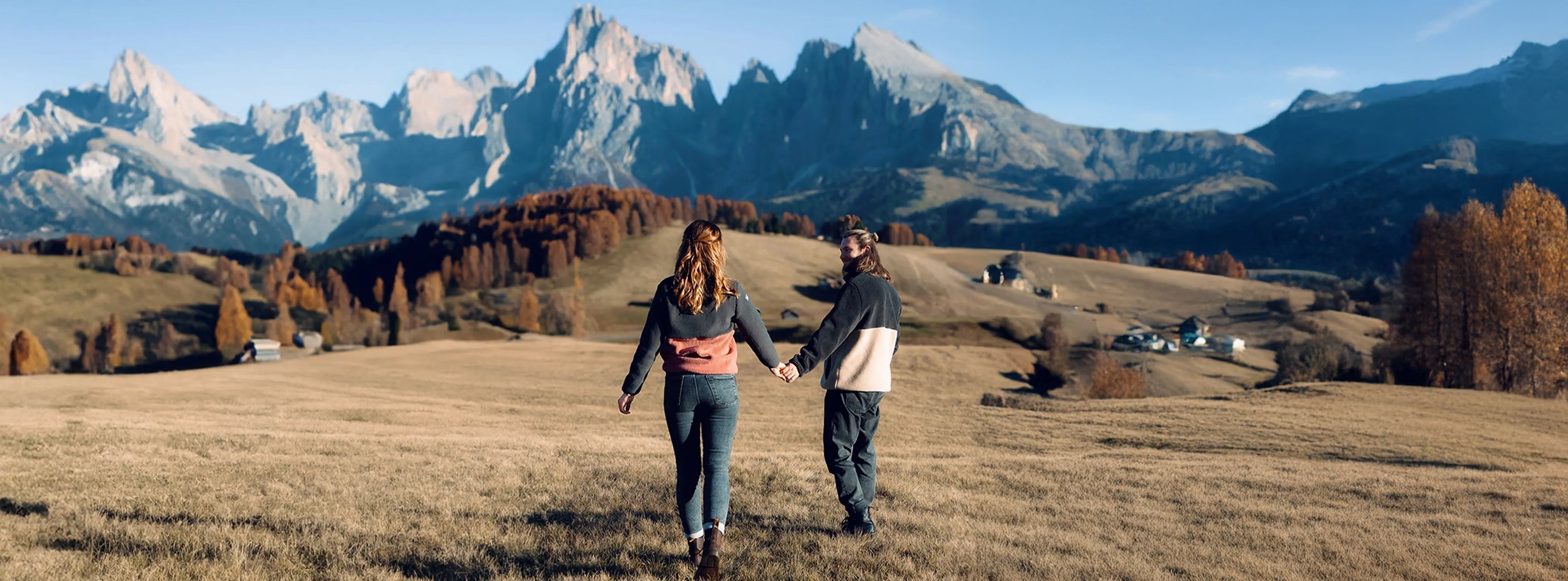 Paar hält Händchen und spaziert durch eine herbstliche Berglandschaft mit weitem Blick auf die Dolomiten im Hintergrund.