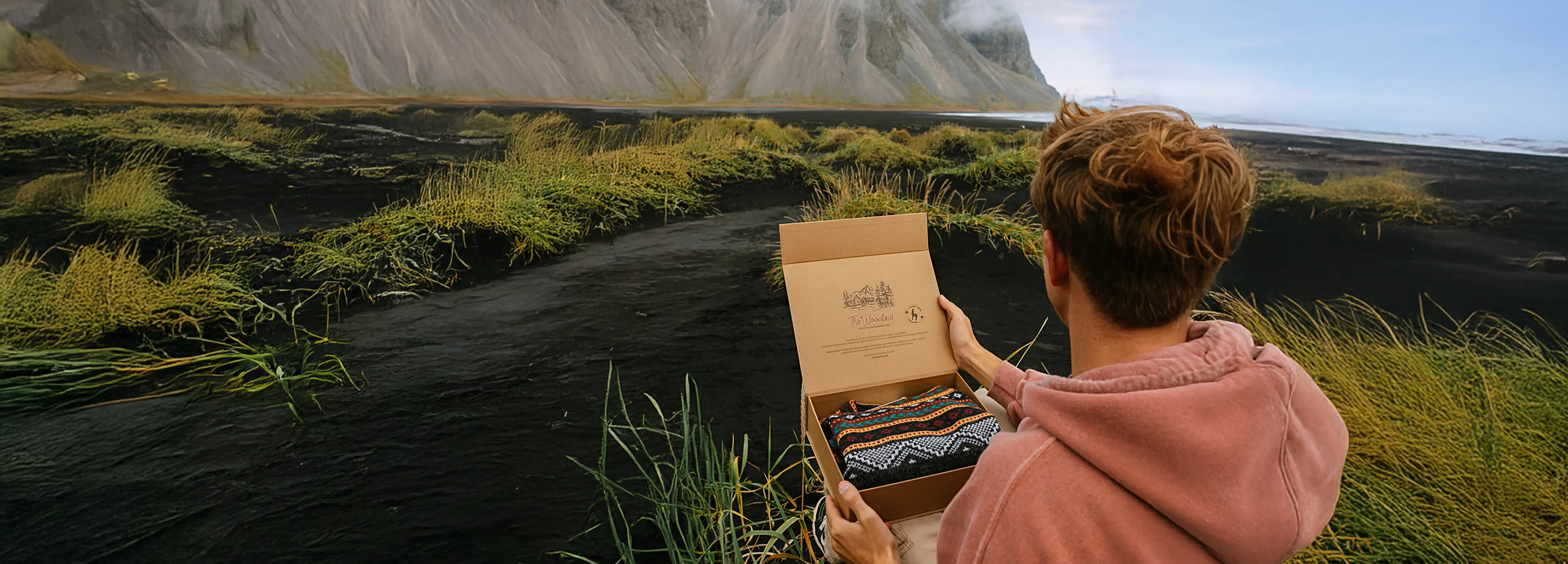 Ein Mann sitzt in der Natur und hält eine Geschenkbox mit einem Alpakapullover darin in der Hand.
