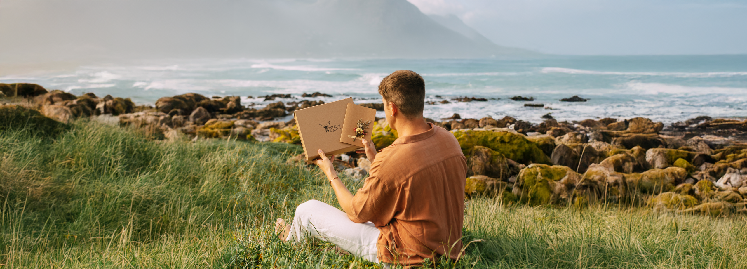 Ein Mann sitzt auf einer Wiese am Meer und hält eine elegante Geschenkbox von Timber&John sowie eine Grußkarte mit echten Blumen in den Händen.