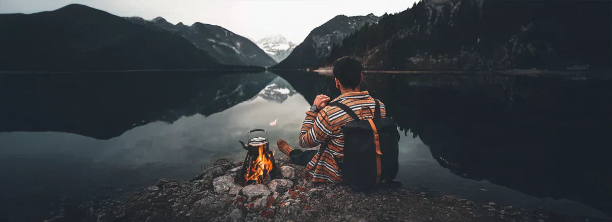 Ein Mann sitzt am Ufer eines Sees neben einem kleinen Lagerfeuer, auf dem eine Kanne mit heißem Wasser steht.