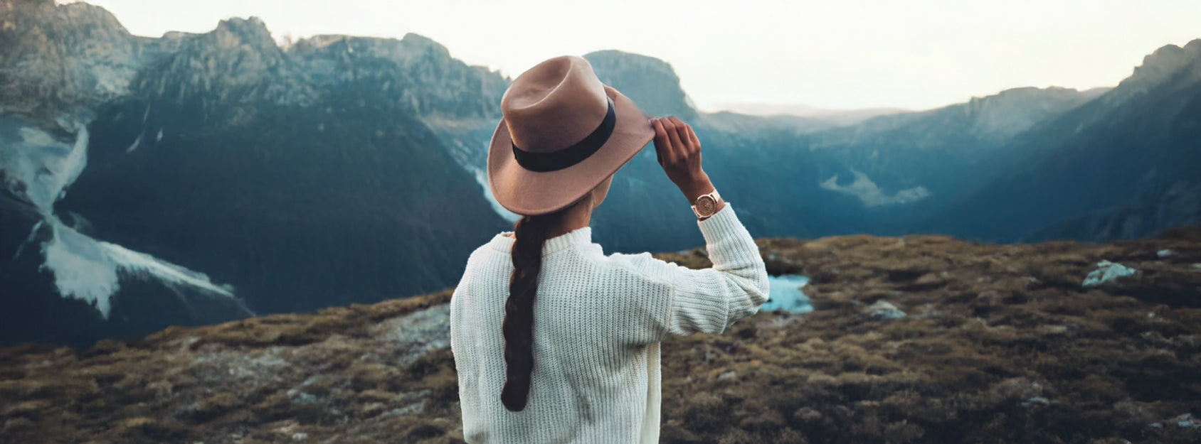 Frau steht auf einem Berggipfel mit Hut und genießt die Aussicht. Am Handgelenk trägt sie eine elegante roséfarbene Damenuhr mit Holzziffernblatt.
